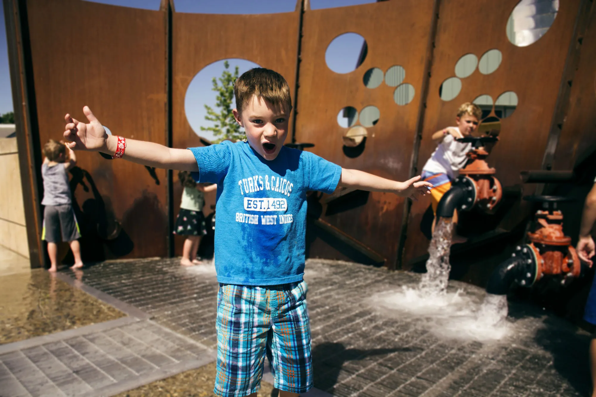 A boy with outstretched arms and a big smile at a water play area while other children play behind him