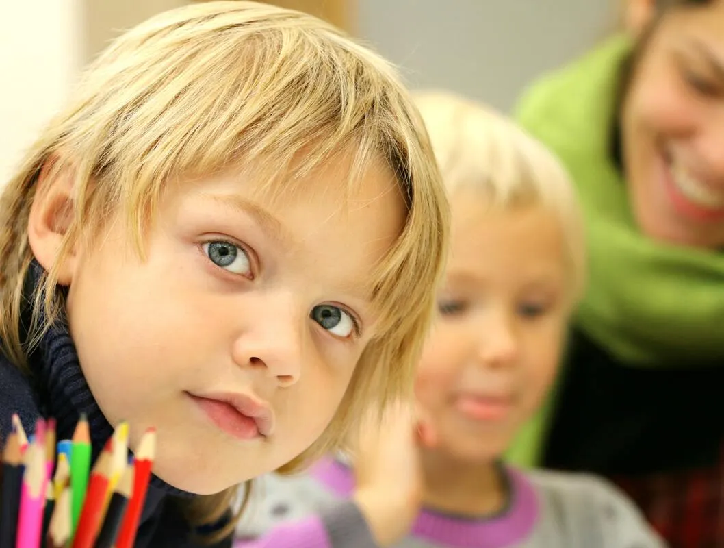 A child with colored pencils in a classroom, with a teacher smiling behind them