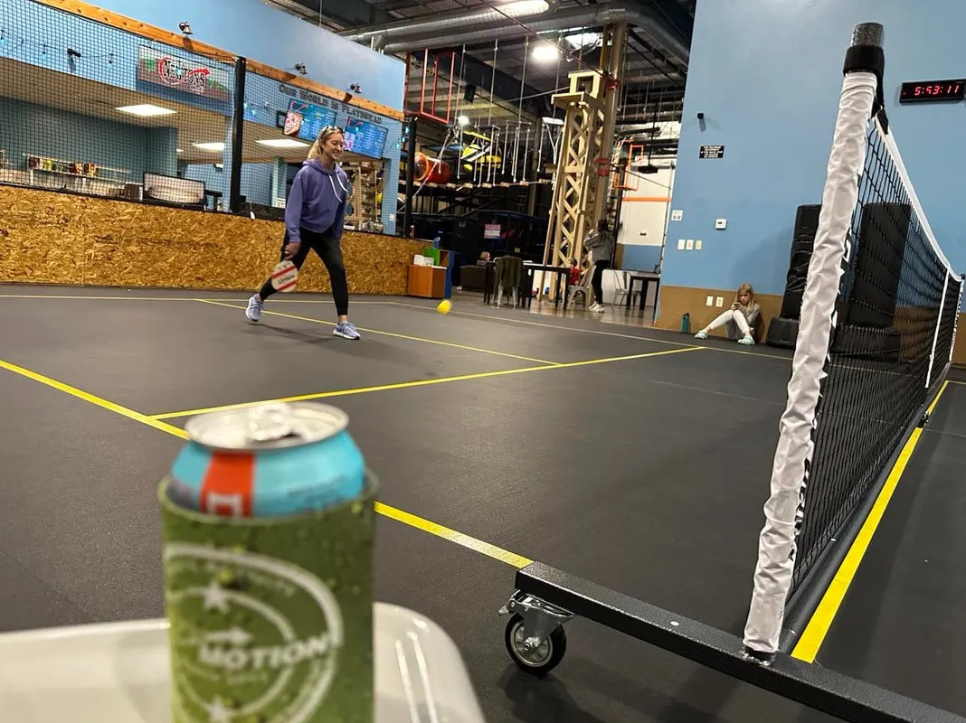 A player tracks a pickleball mid-air on the indoor court, with the ropes course behind